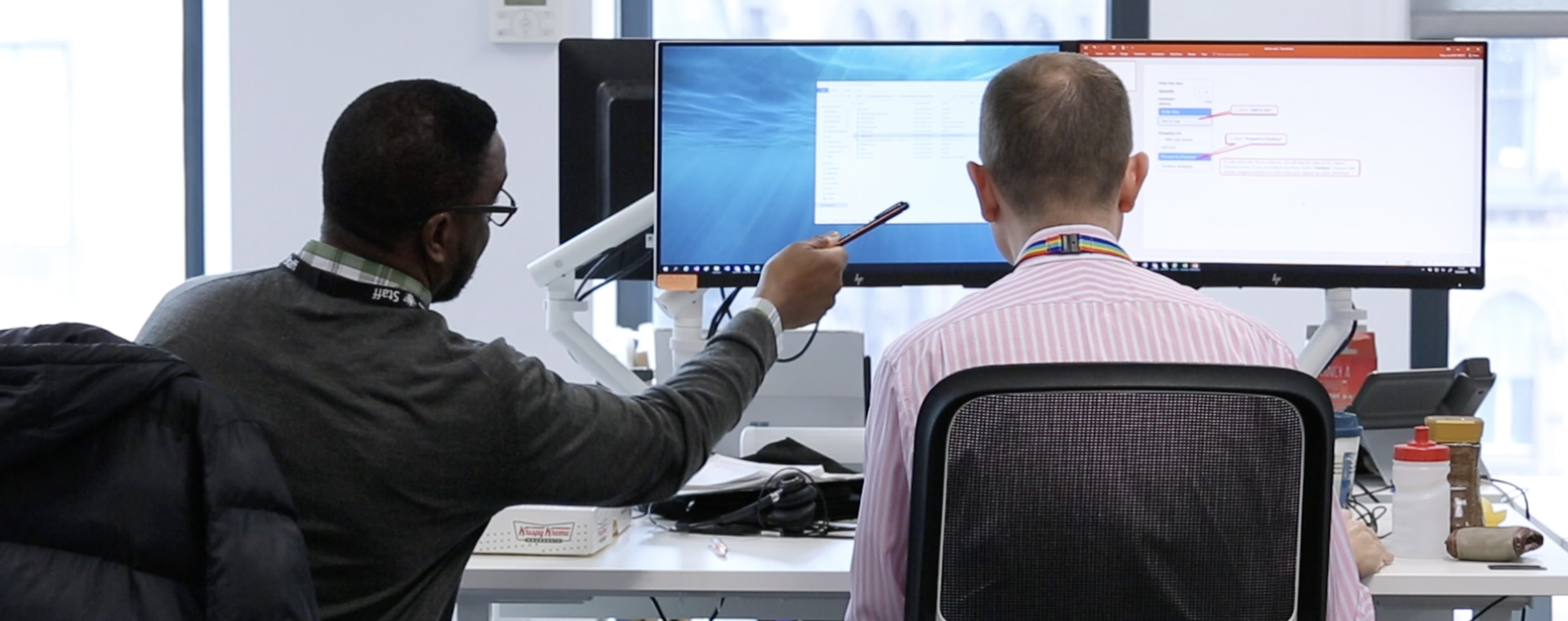 Two people facing away from the camera, seated in front of large computer screens.